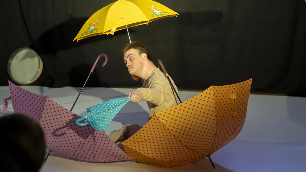 Ein Mann mit Behinderung auf einer Bühne. Er hält einen aufgespannten gelben Regenschirm in der Hand. Vor ihm liegen zwei weitere aufgespannte Regenschirme.