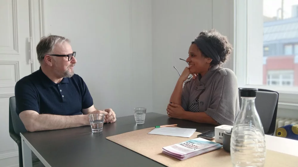 An einem Besprechungstisch sitzen sich die Journalistin Elisabeth Wellershaus und Holger Bergmann, Geschäftsführer des Fonds Darstellende Künste, in einem Gespräch vertieft, gegenüber.