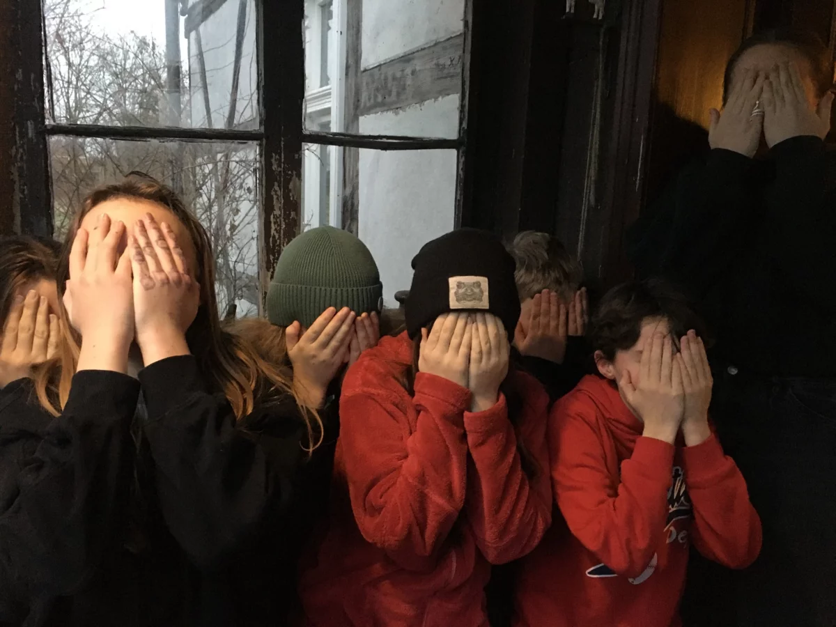 A group of children have positioned themselves in front of a window. They hold their hands in front of their eyes.