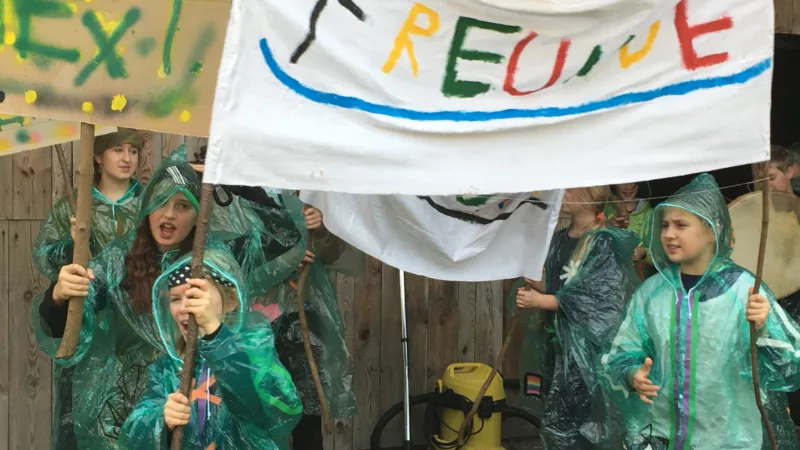 A group of children in green rain ponchos made of thin plastic demonstrate, two children hold a banner with the word Friends written on it in colorful letters.