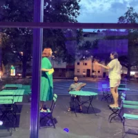 This picture shows three people in front of a bar, standing on chairs and interacting with each other.