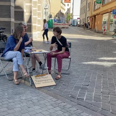 Drei Menschen sitzen an einem Tisch und unterhalten sich. Sie befinden sich auf dem Martkplatz in Halle/Saale.