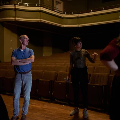 Four people stand in a circle in an empty theater space. They are talking in front of the audience.