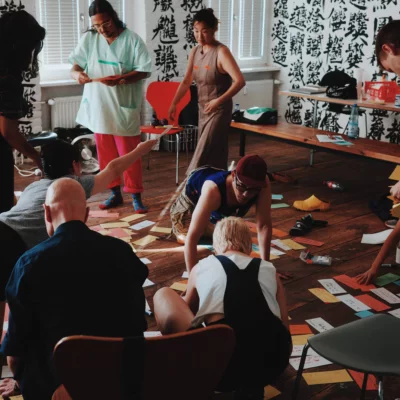 A larger group of people is in a room with small pieces of paper on the floor and other working materials. They enter into exchange with each other.