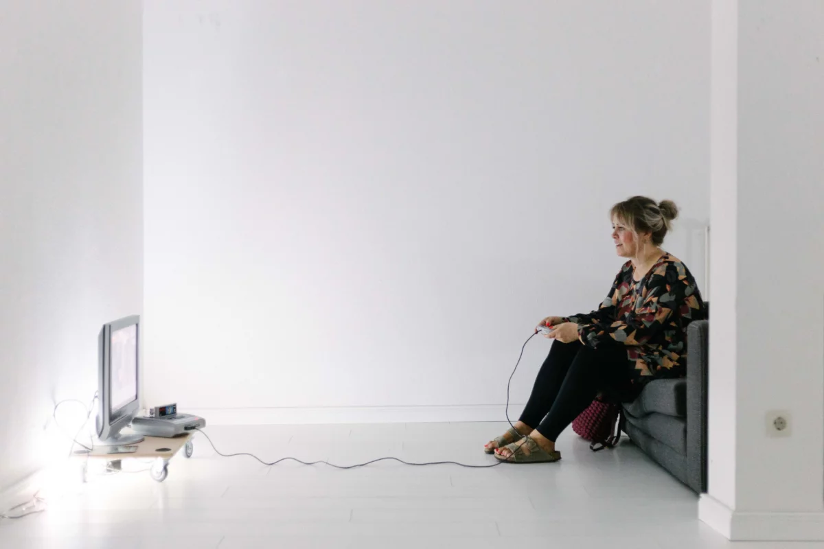 A person is sitting on a chair in a white bare room, playing on a console and looking at a screen.