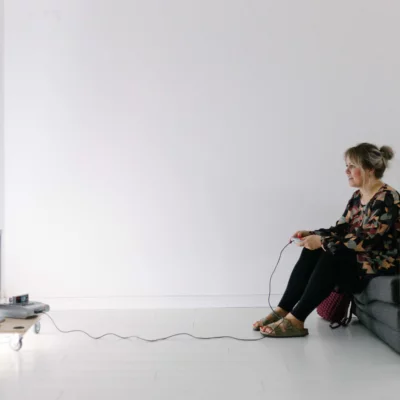 A person is sitting on a chair in a white bare room, playing on a console and looking at a screen.