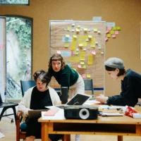 Three people are between work materials in a room surrounded by post-its, a projector and flip charts.