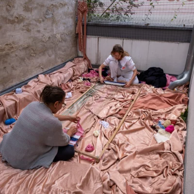 Two people sit on a large pink cloth and work on a wooden frame.