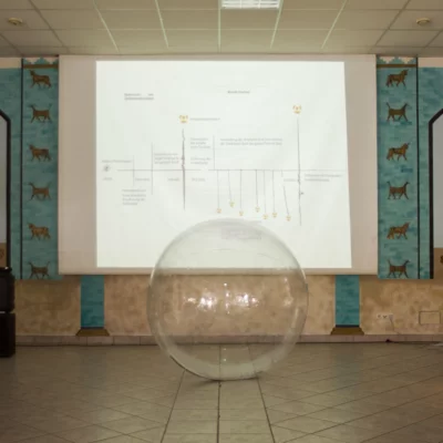 A large transparent plastic ball stands in front of a screen.
