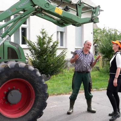 A woman and a man stand in front of a tractor and laugh.