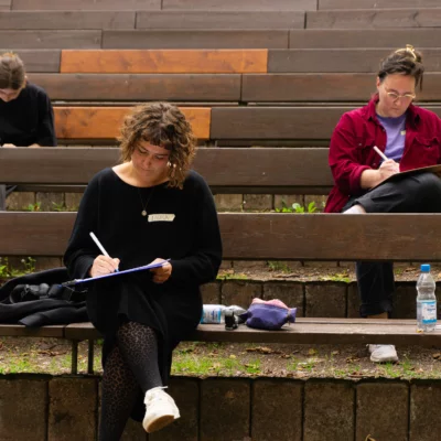 Three people sit staggered on wooden benches and write something down.
