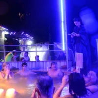 A group of people in swimwear are sitting in an outdoor pool. It is dark. A bright blue fluorescent tube provides illumination. A person in a bathrobe stands at the edge of the pool, holding a piece of paper from which they are reading.