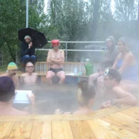 People in swimwear, some wearing caps, sit in an outdoor pool. Steam from the hot water hangs in the air. One person is holding a piece of paper and giving a lecture. The others listen