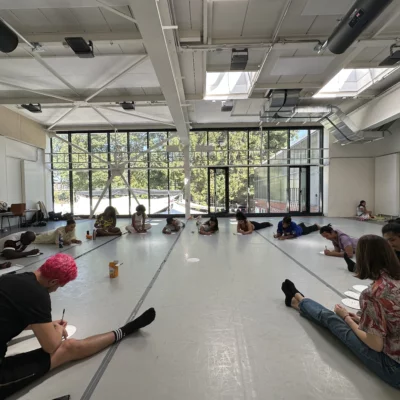 Many people sit together in a circle on the floor in a dance studio.