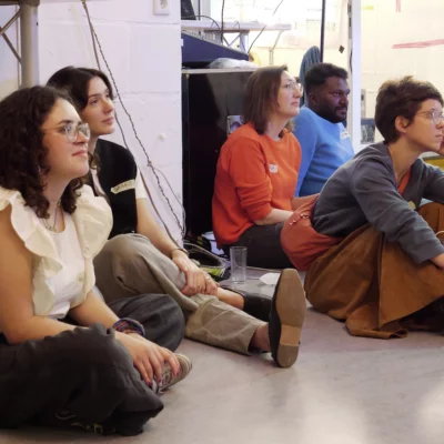A group of people sits on the floor and listens with interest to other people.