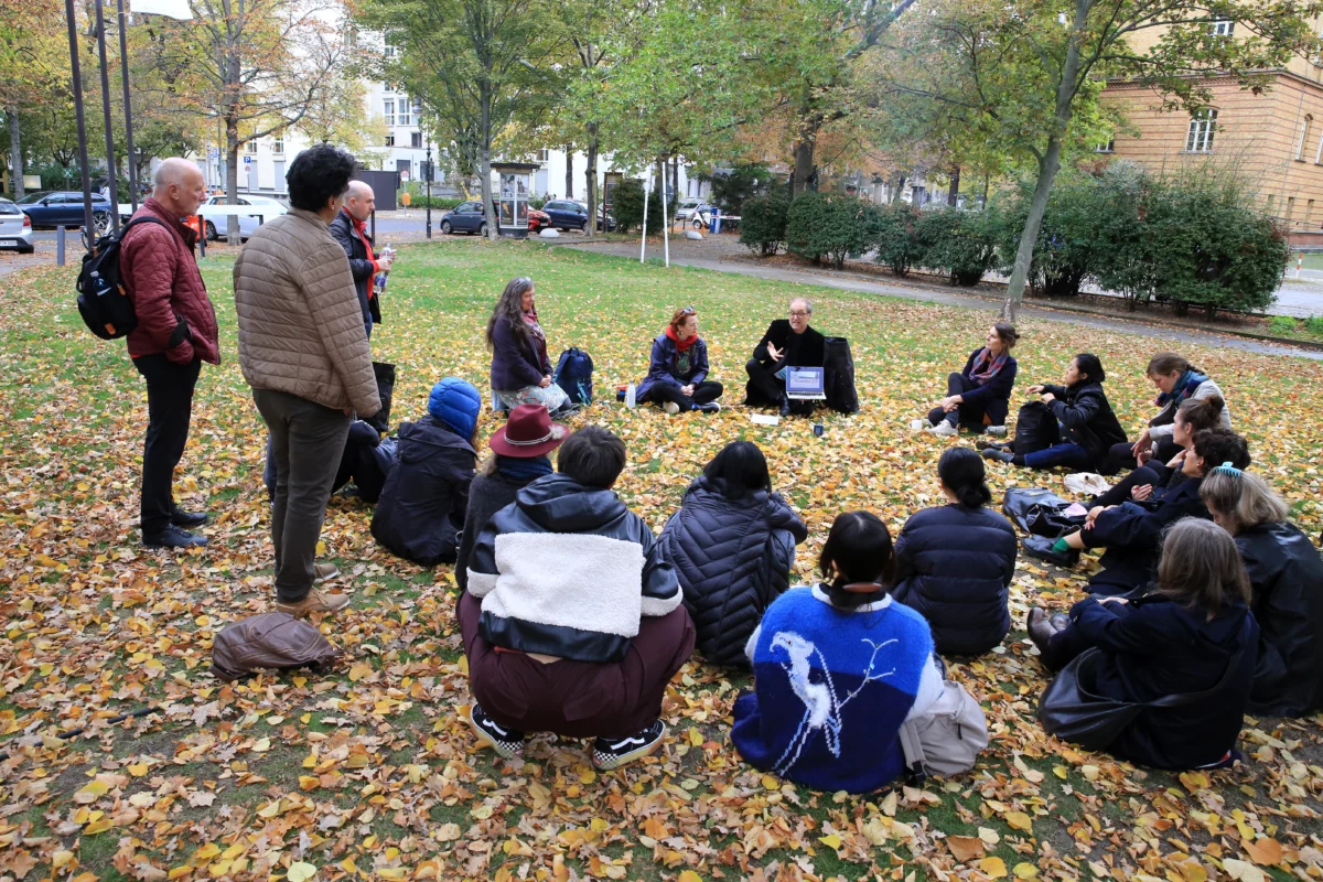 On a lawn covered with autumn leaves, about 20 people sit in a circle and listen to a speaker who holds an open tablet in one hand. Three people stand listening at the edge.