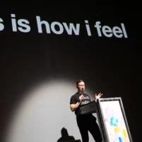 The artist Jörg Albrecht stands at the lectern and gives his impulse talk on stage. Behind him is a black screen on which the words "this is how i feel" are projected.