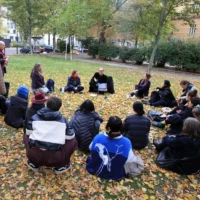 Auf einer mit Herbstlaub bedeckten Wiese sitzen rund 20 Menschen im Kreis und hören einem Redner zu, der in einer Hand ein offenes Tablet hält. Drei Menschen stehen lauschend am Rand.