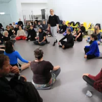 Many people sit cross-legged during a workshop in a circle around choreographer Ben J. Riepe.