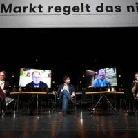 Three people are sitting on the large stage of the Berliner Festspiele for a panel discussion. Between them are two monitors, via which two guests from outside are digitally connected. Above their heads is a banner with the inscription: "The market doesn't regulate it".