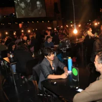 Menschen jeweils zu zweit im Gespräch an Tischen auf der Bühne der Festspiele. Im Hintergrund ist eine Leinwand zu sehen, auf die Gesichter von zwei Gesprächsteilnehmer*innen zu sehen sind.
