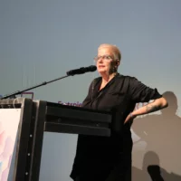 The picture shows the artist and scientist Sibylle Peters. Standing at the lectern, she gives her keynote speech to the audience in the hall.