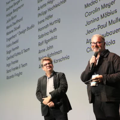 The picture shows the managing director of the Fund, Holger Bergmann, and the artistic director of the Berliner Festpsiele, Matthias Pees, in front of a screen on stage as they welcome the audience in the hall.