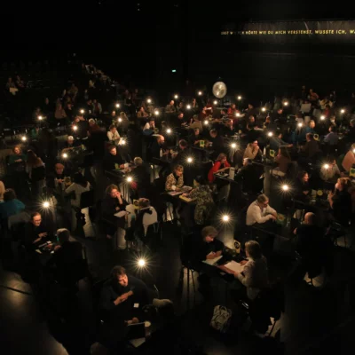 In the darkened stage area, people sit at around 50 tables, two by two, engrossed in conversation. Above each table, a dimly lit bulb provides light in the dark.