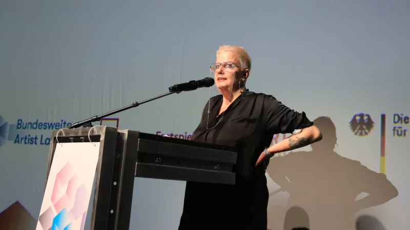 The picture shows the artist and scientist Sibylle Peters. Standing at the lectern, she gives her keynote speech to the audience in the hall.