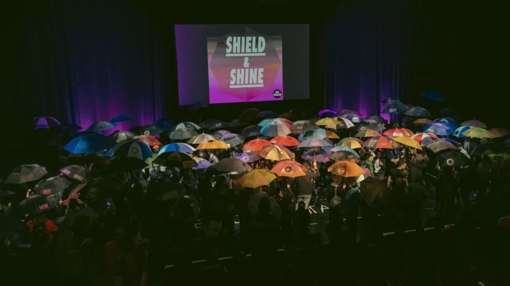 There are many people on a stage, all holding a stretched umbrella in their hands. On a projection surface in the background is written: Shield and Shine.
