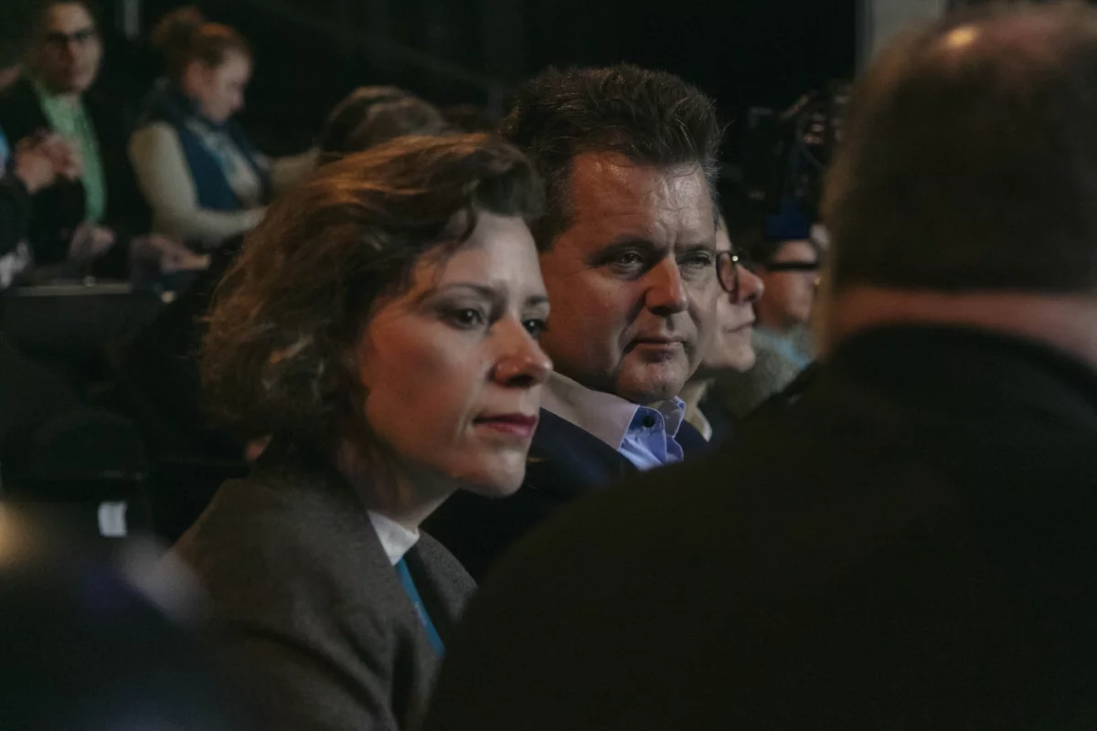 In the left half of the picture is Aniko Glogowski-Merten. She has brown chin-length hair. Jürgen Dusel is sitting next to her. He is wearing a white shirt and a dark jacket. He has short dark hair. Both are looking in the direction of a person whose outline appears in the blur in the front right half of the picture.