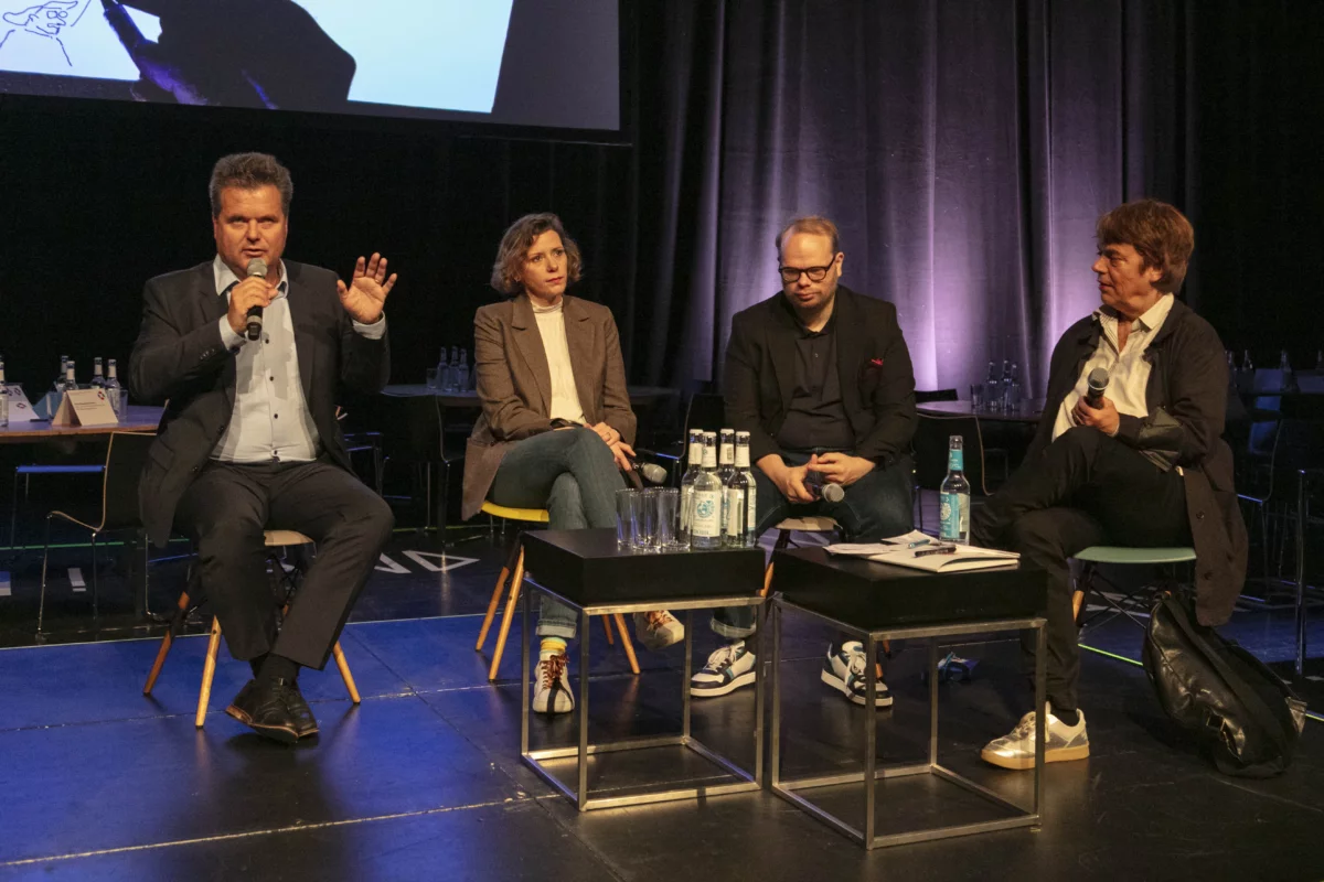 Four people are sitting on a stage in a panel discussion. On the right, a man in a suit speaking into a microphone. To his left, a woman, a man and another woman listen intently.