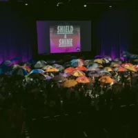 There are many people on a stage, all holding a stretched umbrella in their hands. On a projection surface in the background is written: Shield and Shine.