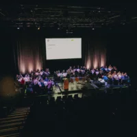 On a stage, several round tables are arranged, at which people are sitting, talking to each other. In the background hangs a projection screen on which "Table Talks" is written.