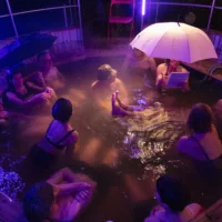 People sit in a heated outdoor pool and talk to each other. One person is holding a stretched umbrella. It is evening and dark. Steam from the warm water hangs in the air.