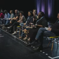 Zehn Menschen sitzen aufgereiht auf einer Bühne. Sie halten Zettel in der Hand. Eine Schwarze Frau spricht in ein Mikrofon. Auf vielen Gesichtern ist ein Lächeln zu sehen.