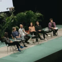 Six people sit lined up on a stage in a panel discussion. Two of them hold microphones in their hands. Behind them are large houseplants.