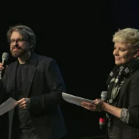 On the left, a man with a full beard and medium-length hair in a dark suit holding a microphone and speaking. On the right, a person with short blond hair and scarf giving applause.