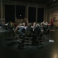 Large round tables are spread out in a darkly lit room. The tables are more brightly lit. At them sit people who are engrossed in conversation.