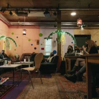 In a room, tables, sofas and chairs are distributed in a cozy atmosphere. People with FFP2 masks and sufficient distance sit everywhere and talk to each other. The walls are yellow and pink. There are two self-made palm trees illuminated with fairy lights.