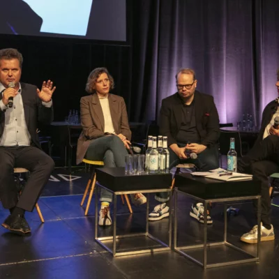 Four people are sitting on a stage in a panel discussion. On the right, a man in a suit speaking into a microphone. To his left, a woman, a man and another woman listen intently.