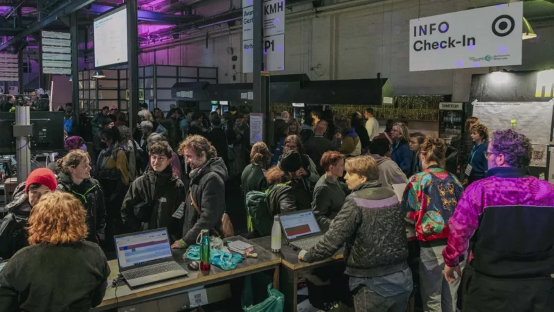 There are many people in the foyer at Kampnagel. People are standing at laptops at a counter. Above them hangs a sign with the inscription "Info / Check-In.