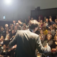 Im Vordergrund ist Dr. Günter Winands während seines Vortrags in einer Rückansicht zu sehen. Im Bildhintergrund das Publikum.