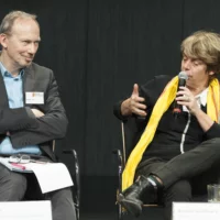 Amelie Deuflhard expresses her thoughts at a panel discussion, microphone in hand. Martin Eifler (BKM) sits next to her and listens to her remarks with folded arms.