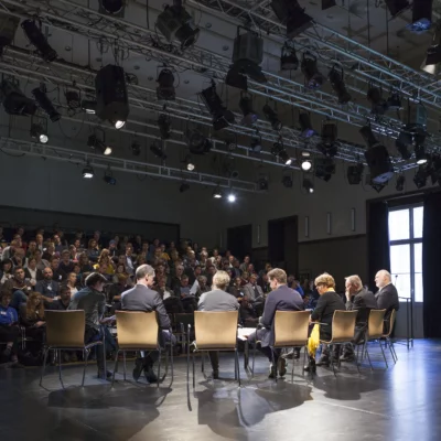 Sieben Podiumsgäste sitzen im Halbkreis zum Gespräch auf der Bühne der Sophiensaele. Das Bild zeigt sie in einer Rückansicht: vor ihnen sind volle Ränge mit Publikum zu sehen.