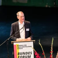 Portrait of Hans Heinrich Bethge at the lectern, on which a poster of the event is attached. He speaks into the microphone, holding index cards in his hand. Next to the lectern, red gladioli can be seen in the otherwise dark stage space in the background.