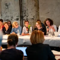 Several people sit at a long table with a white paper tablecloth. Annemie Vanackere speaks into a microphone and underlines her contribution gesturally.