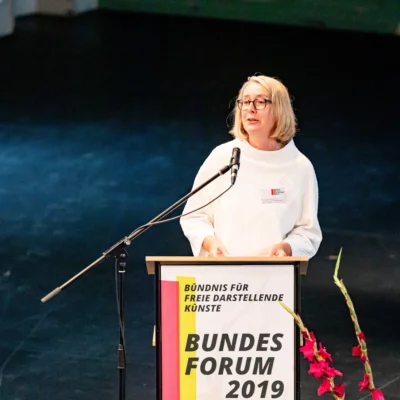 Portrait of Dr. Sigrid Bias-Engels (BKM) at the lectern, to which a poster of the event is attached. She speaks into the microphone, her eyes directed forward to a non-visible audience. Next to her are two gladioli in a vase.