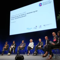 Nine people sit side by side on chairs on the podium in a circle of chairs for the pdoium discussion. Above them is a large screen on which the title "Opening Federal Forum 2021: Positions from politics on changing the art and cultural landscape" can be read.