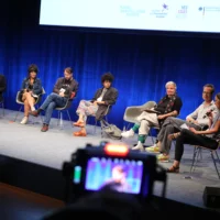 Seven people sit in a circle of chairs for a panel discussion on stage.